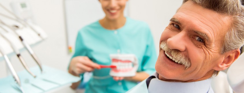 Older gentleman patient in dental practice with hygenist shoing how to brush dentures